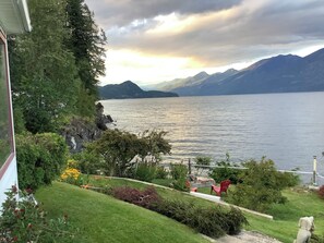 A lakeside view with mountains in the background, a grassy area with a pathway, and a chair. - Vacation Home For Rent - Ainsworth BC (Ainsworth)