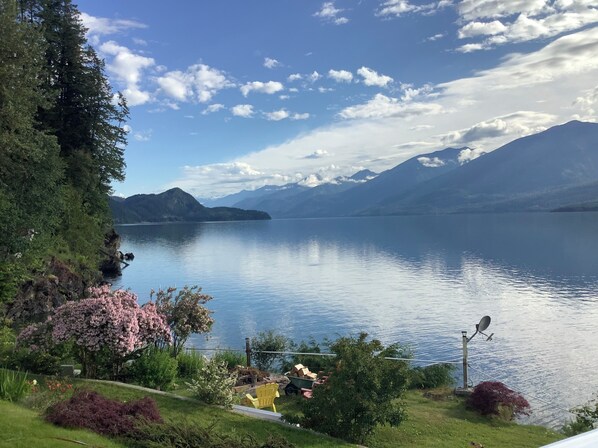 A serene lakeside view with mountains in the background, a clear sky, and a well-maintained garden area with a yellow chair and a satellite dish. - Vacation Home For Rent - Ainsworth BC (Ainsworth)