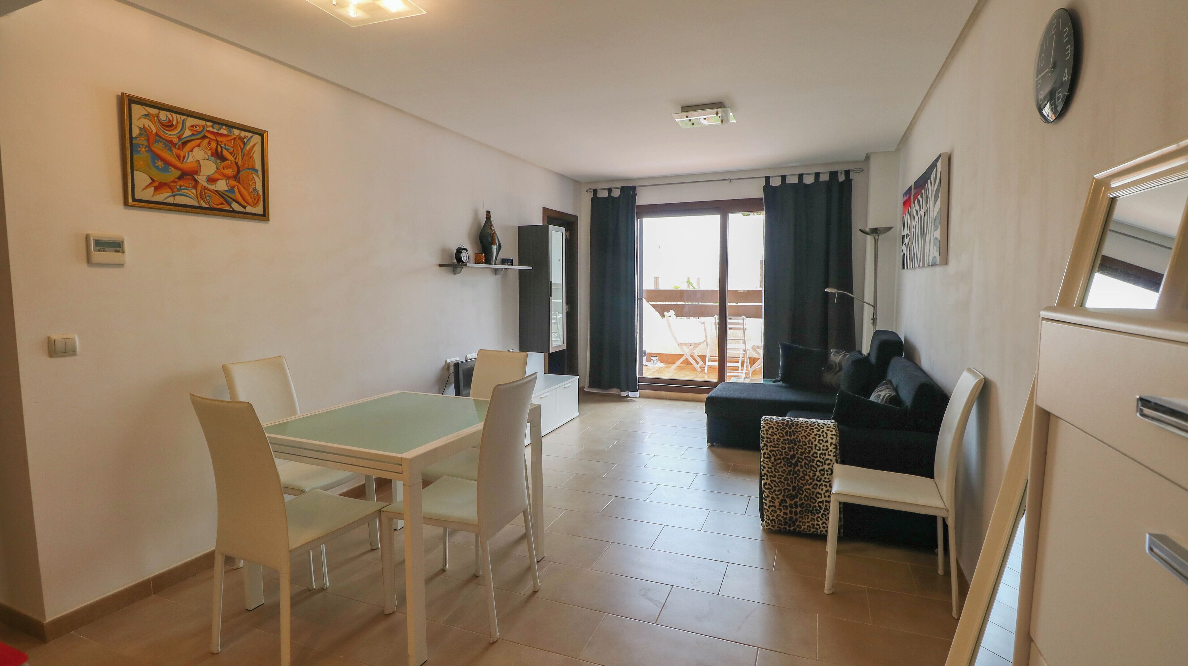 A modern living room with a sofa, a dining table with chairs, a kitchen area, and a balcony with a table and chairs.