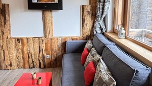 A cozy living room with a wooden accent wall, a flat-screen TV mounted on it, a red coffee table, and a grey sofa with patterned cushions.