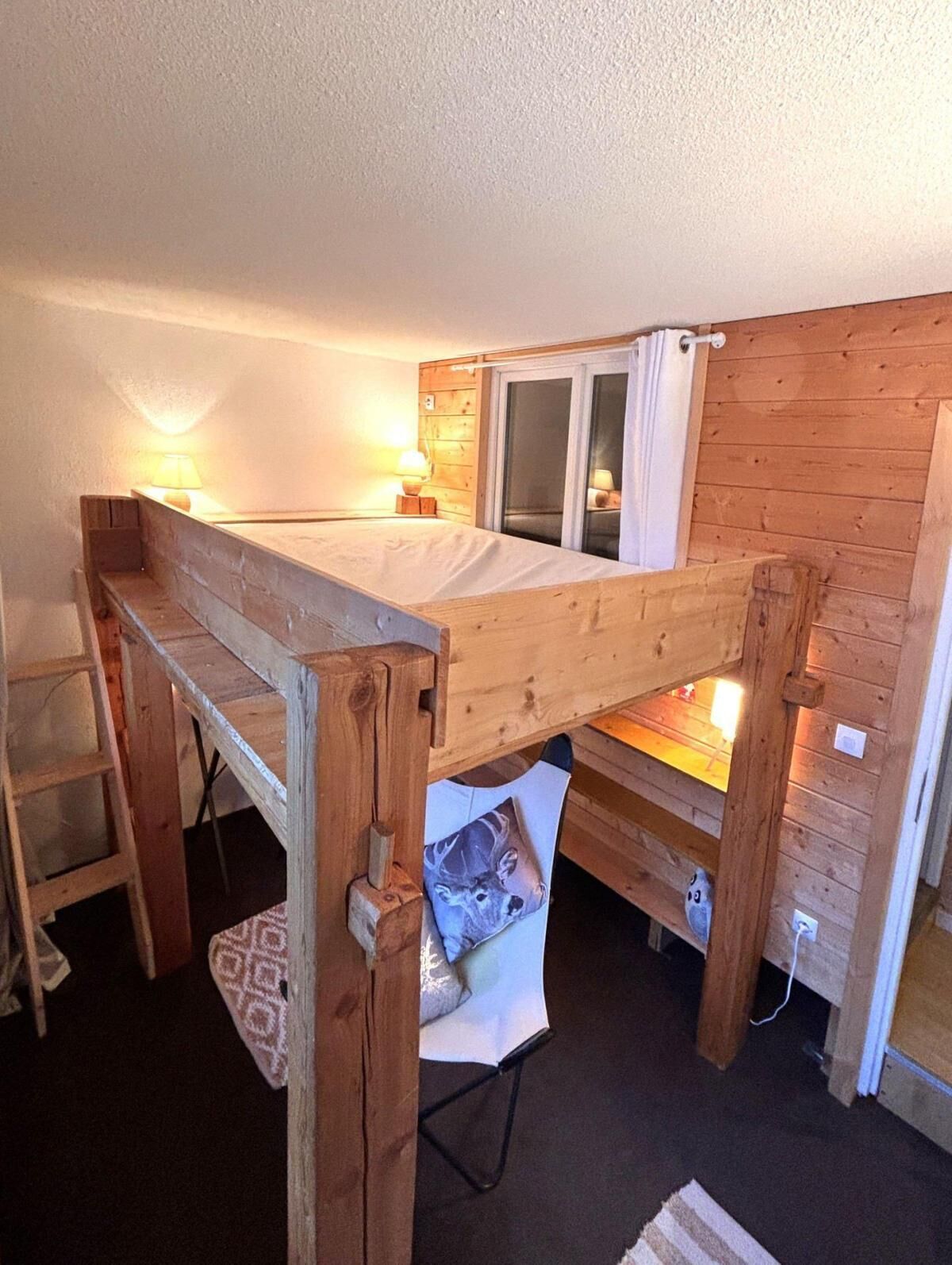 A wooden bunk bed with a ladder, a window with curtains, and a bedside lamp.