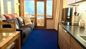 Une pièce de taille modeste comprenant un canapé, une table en bois, un coin cuisine avec des placards et une grande fenêtre donnant sur les montagnes.