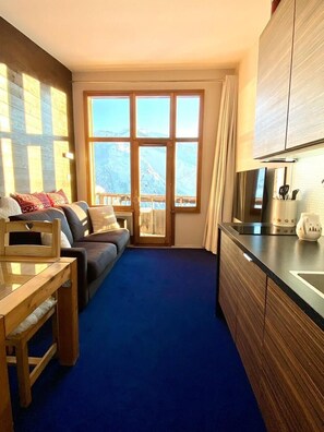 A compact room with a sofa, a wooden table, a kitchen area with cabinets, and a large window with a view of mountains. - 200m from resort center, 7th floor, balcony, tv, ski locker, 26m², Avoriaz (Avoriaz)