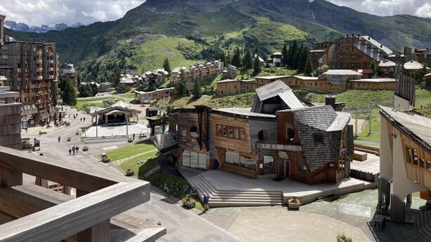Un pueblo montañoso con edificios tradicionales de madera, una plaza central y vistas a montañas distantes.