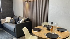 A modern living room with a grey sofa, a round wooden dining table set with plates, glasses, and cutlery, and a vertical wooden panel wall.