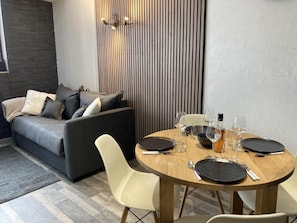 Ein modernes Wohnzimmer mit einem grauen Sofa, einem runden Holzesstisch gedeckt mit Tellern, Gläsern und Besteck, sowie einer Wandpaneele aus Holz.