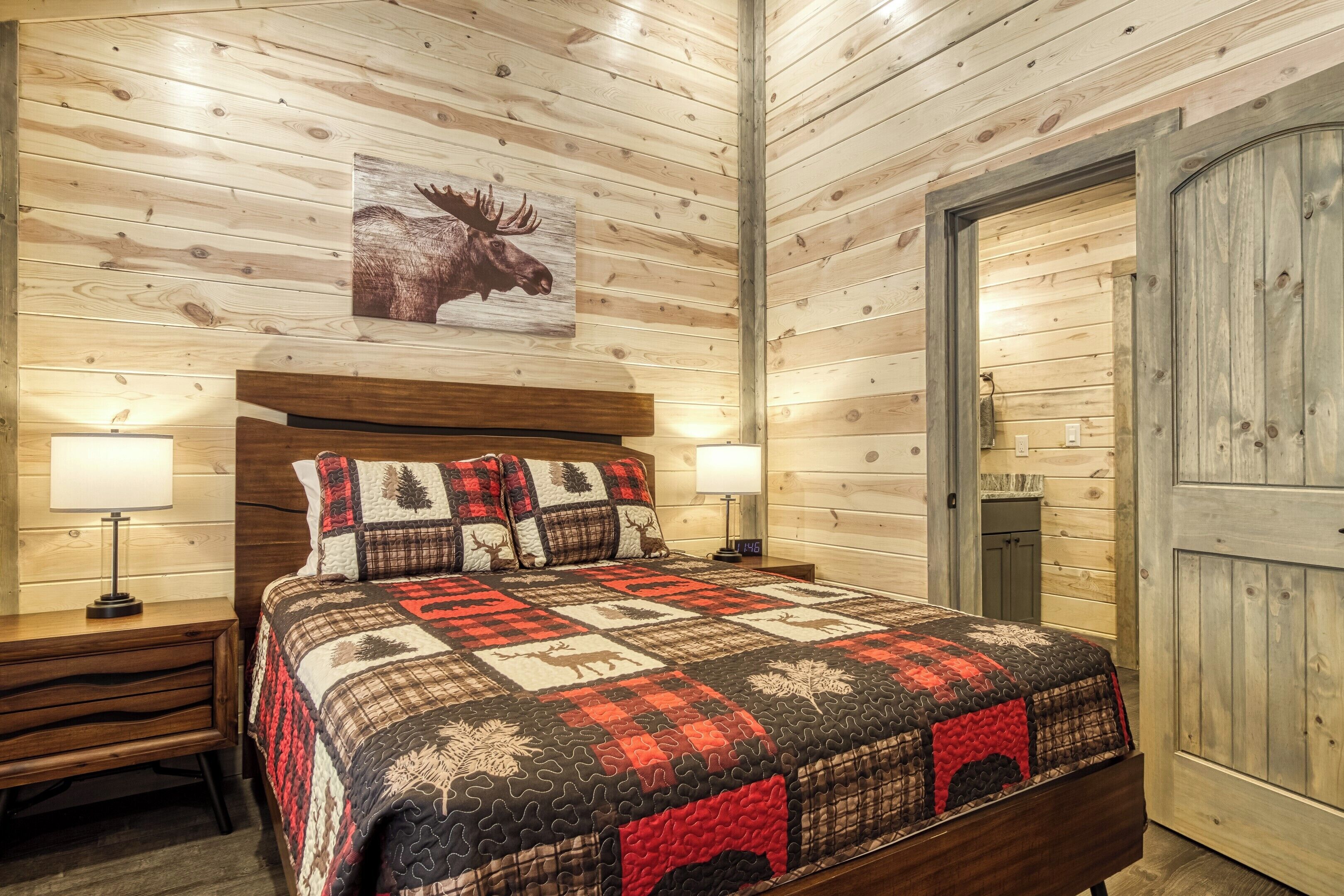 Une chambre confortable avec un plafond en bois, un grand lit recouvert d’une courtepointe, deux tables de chevet avec des lampes et une porte sur le côté droit.