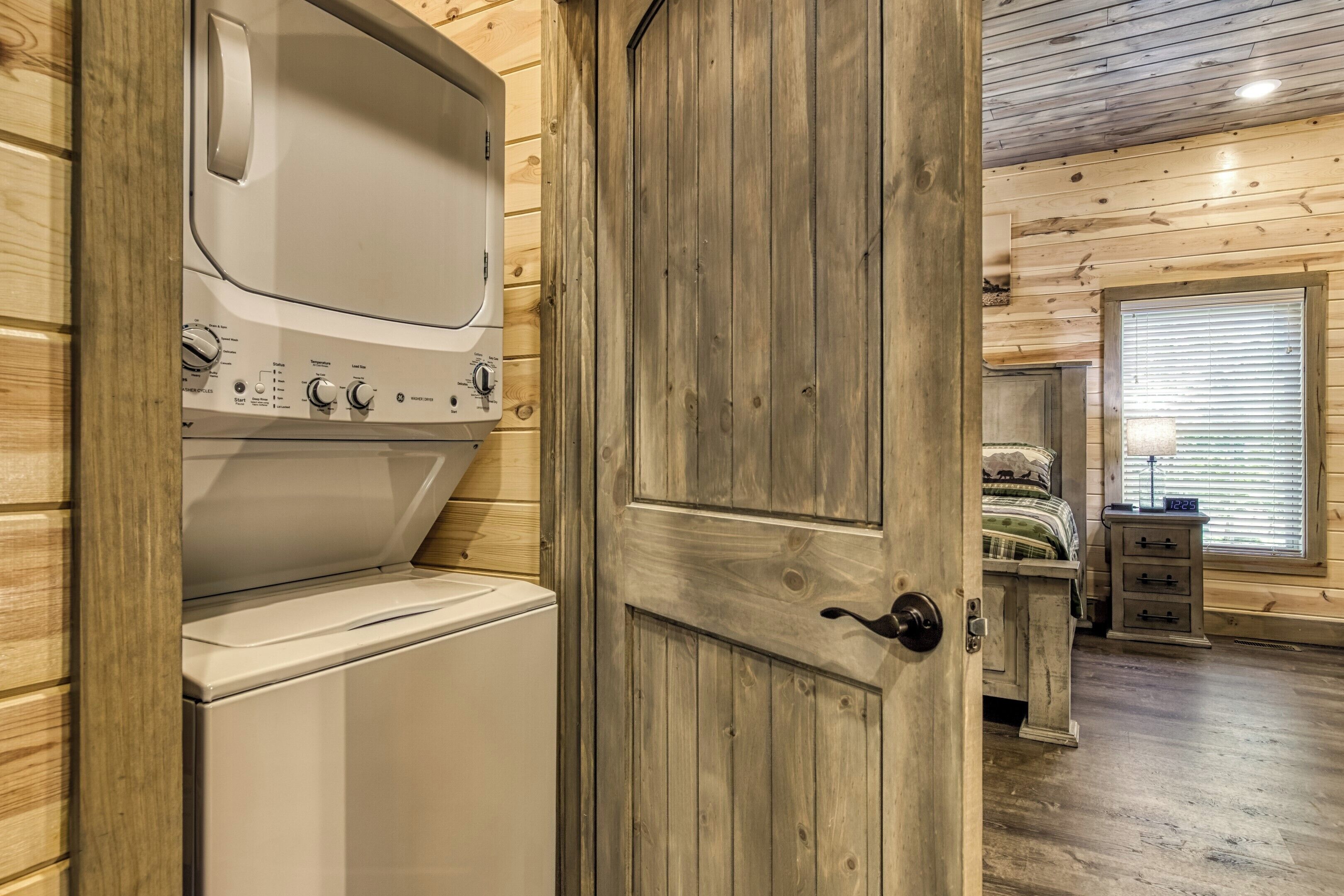 A laundry room with a washer and dryer, wooden walls, a bed with a nightstand, and a window with blinds.