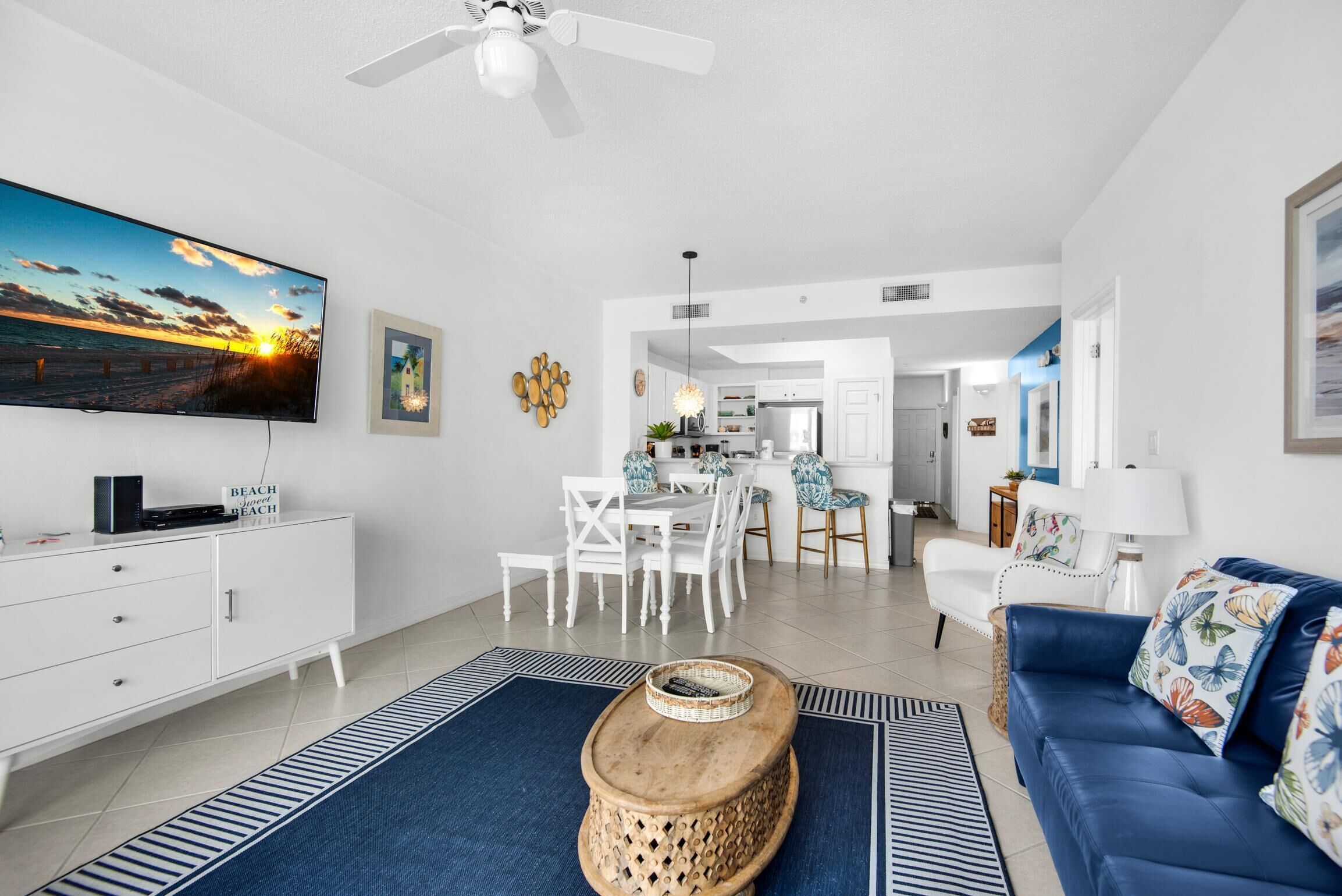 A modern living room with a flat-screen TV, a wooden coffee table, a blue sofa, and a dining area with white chairs.