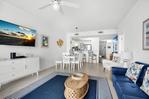 A modern living room with a flat-screen TV, a wooden coffee table, a blue sofa, and a dining area with white chairs. - Summerwind West 1203 (Navarre)
