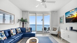 A living room with a blue sofa, a TV, and a balcony with a view of the ocean. - Summerwind West 1203 (Navarre)