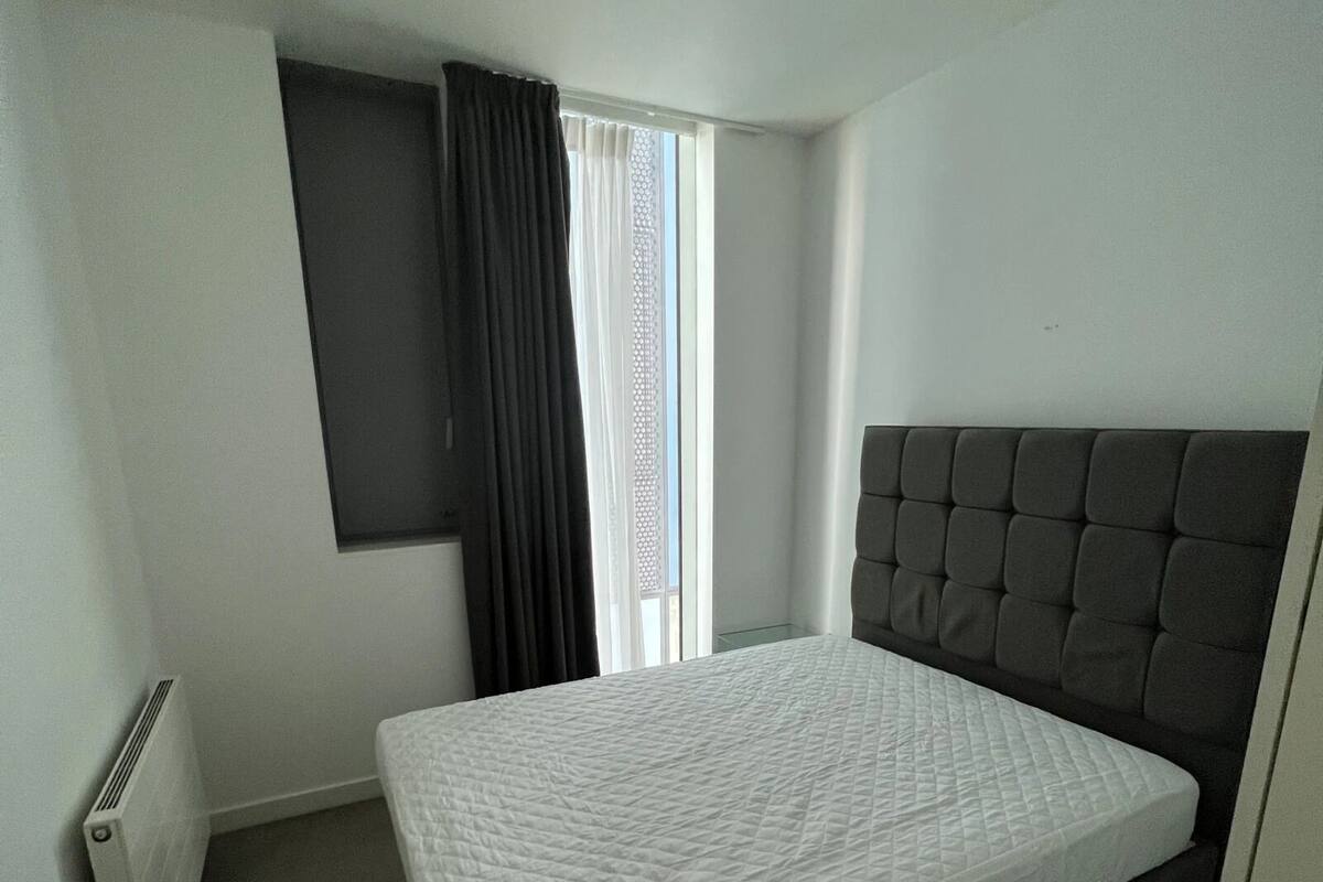 A single bed with a quilted mattress in a room with a radiator, a window with curtains, and a plain white wall.