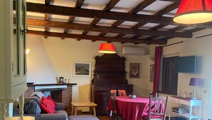 Ein gemütliches Zimmer mit Kamin, einem Esstisch mit rotem Tischlein und einer Holzbalkendecke.