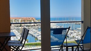 Ein Balkon mit zwei blauen Stühlen, einem Tisch und Blick auf einen Yachthafen mit Booten und Gebäuden im Hintergrund.