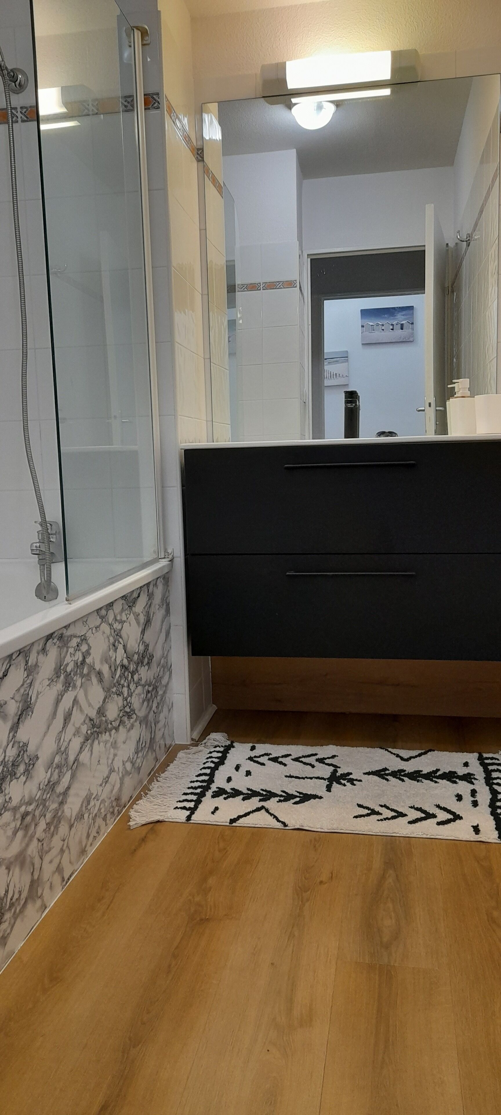 Casa de banho com cabine de chuveiro em vidro, toucador preto com espelho e tapete estampado no chão.