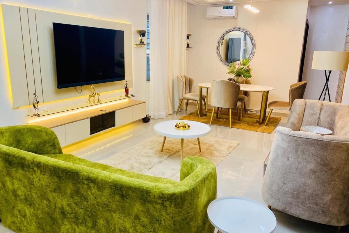 A modern living room with a flat-screen TV, green and beige sofas, a round coffee table, and a dining area with a white table and chairs.