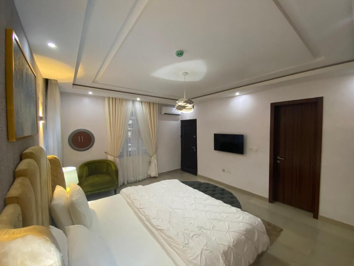 A modern bedroom with a large bed, a green armchair, a TV mounted on the wall, and a door.