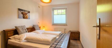 A bedroom with a wooden bed, two bedside tables, a lamp, a radiator, and a window with blinds.