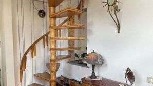 Una scala a chiocciola in legno conduce a un piano superiore, con una scrivania, una lampada, una sedia e un elemento decorativo a parete.