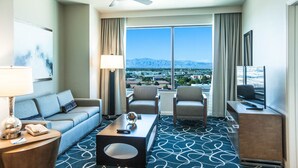 A hotel room with a sofa, two armchairs, a coffee table, a TV, and a large window with a mountain view.