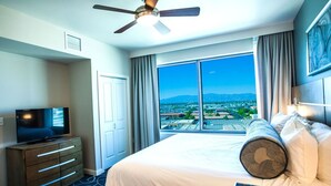A hotel room with a bed, a television, a ceiling fan, and a large window with a city view.