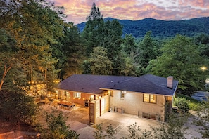 A two-story house with a patio, surrounded by dense forest and hills. - 6BR Asheville House | Hot Tubs, Sauna + Theater (Fairview)