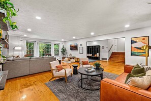 A spacious living room with a large sofa, armchairs, a coffee table, and a staircase leading to another level. - 6BR Asheville House | Hot Tubs, Sauna + Theater (Fairview)