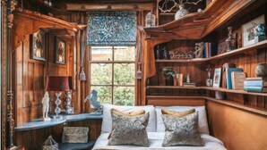 Une pièce chaleureuse avec un décor intérieur en bois, un lit agrémenté d’oreillers à motifs, une fenêtre donnant sur un paysage verdoyant et des étagères remplies de divers objets.