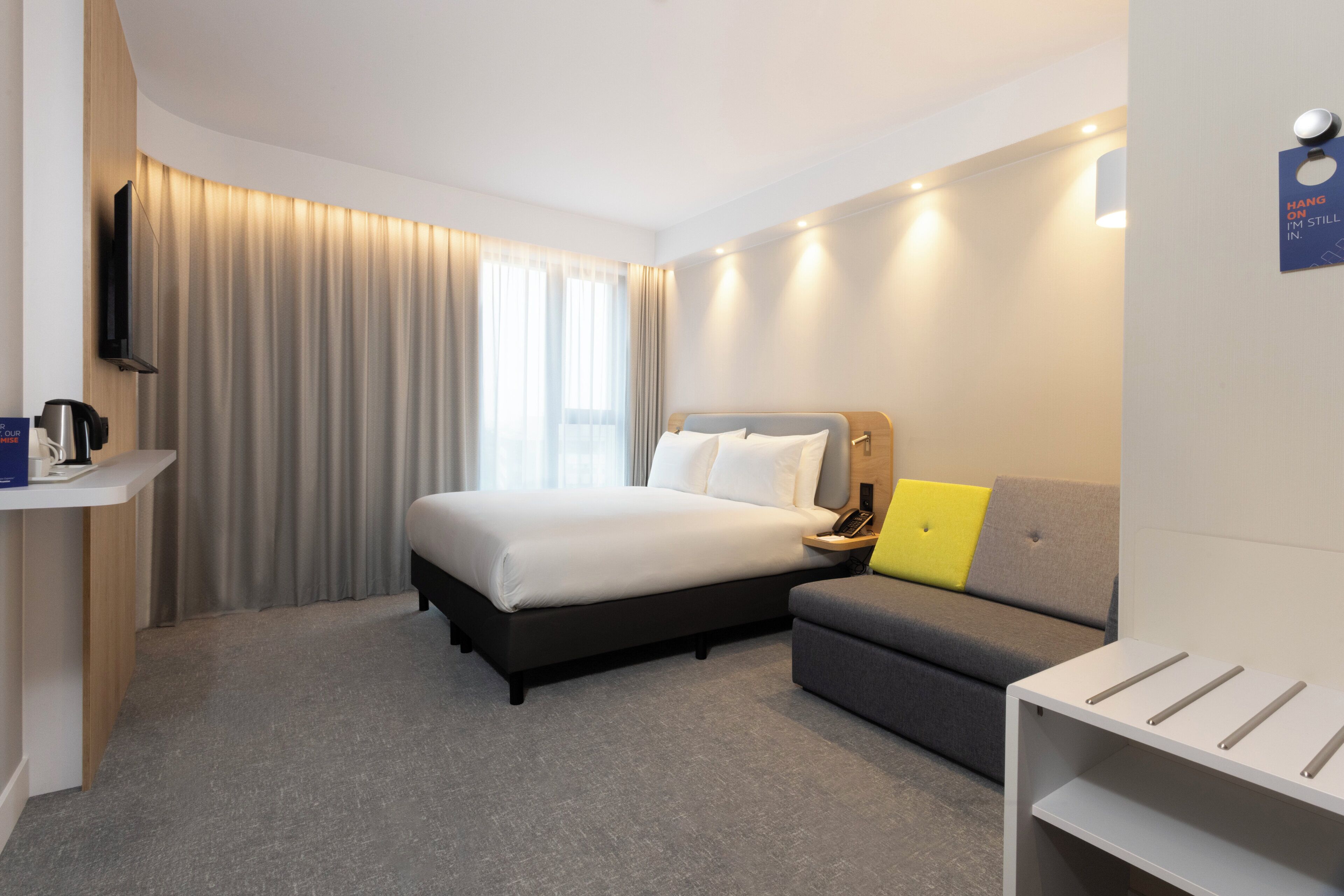 A hotel room with a bed, a sofa, a bedside table, a TV, and a window with curtains.