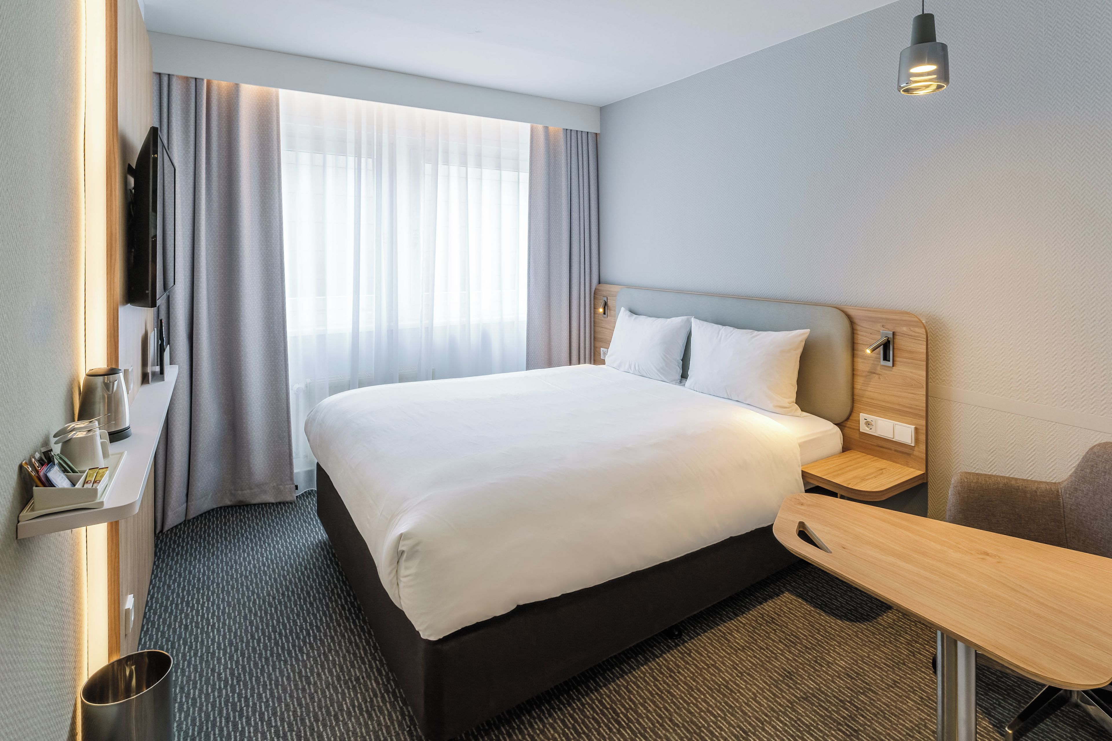 A hotel room with a large bed, a bedside table, a desk, a chair, a TV, and a window with curtains.
