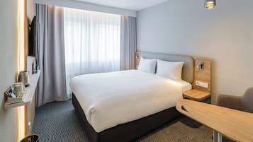 A hotel room with a large bed, a bedside table, a desk, a chair, a TV, and a window with curtains.