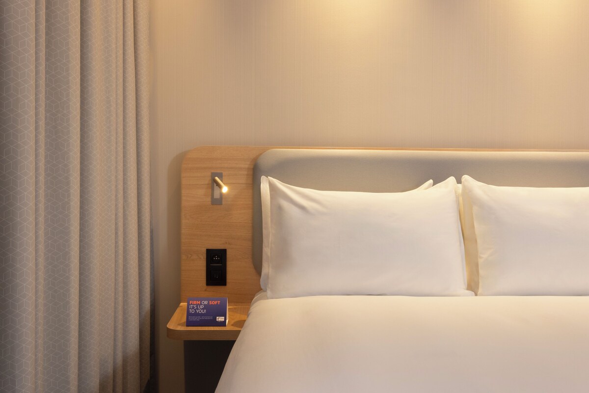 A hotel room with a wooden headboard, a bedside table with a lamp, and a sign indicating room temperature control.