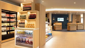 A hotel lobby with a reception desk, seating area, and a variety of food and beverage options including a refrigerated display and shelves stocked with snacks and drinks. - Holiday Inn Express Brussels Airport by IHG (Machelen)