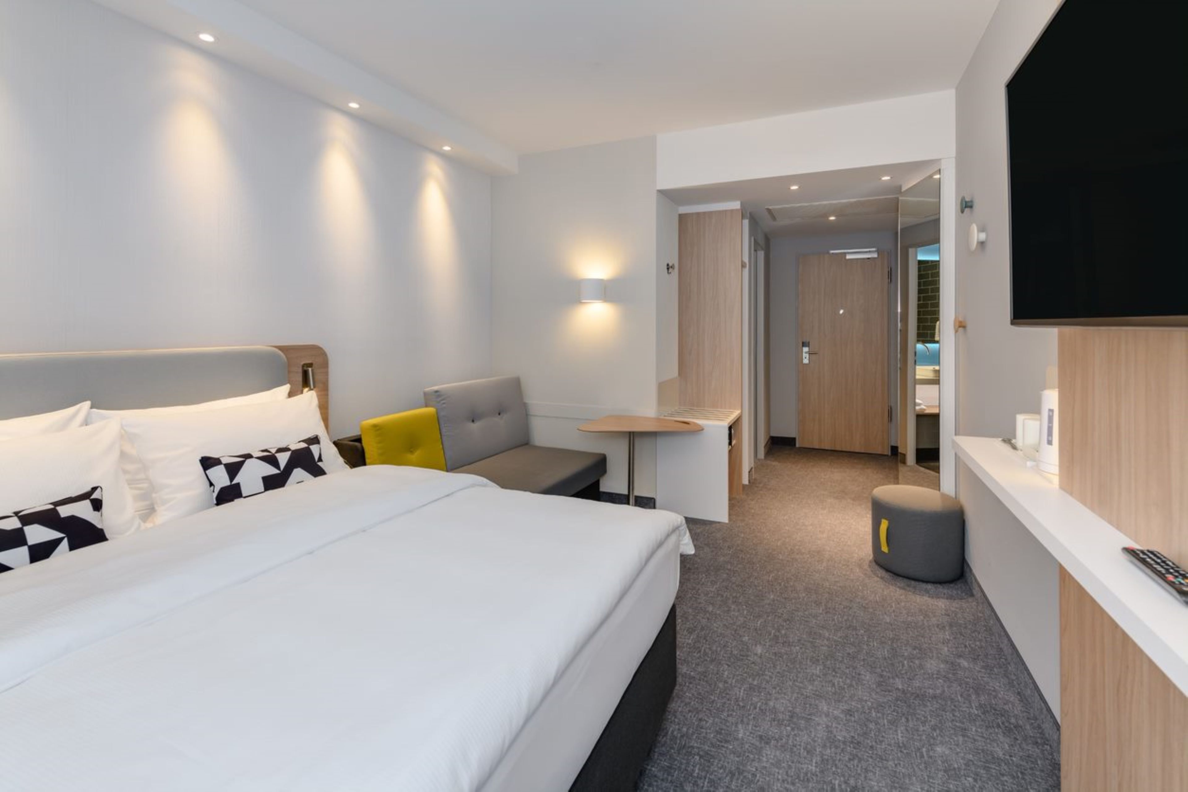 Una habitación de hotel moderna con una cama grande, una pequeña mesa, un sofá, un televisor y un baño visible a través de una puerta abierta.