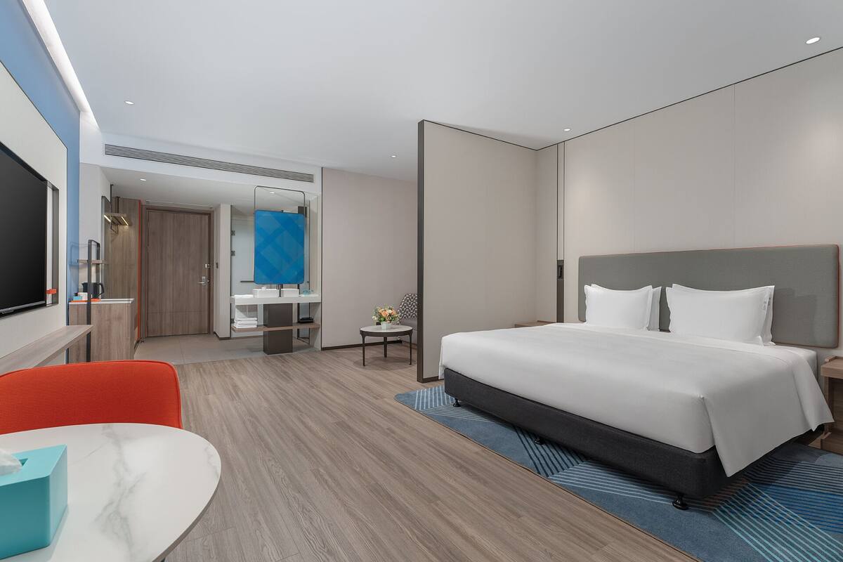 A modern hotel room with a large bed, a desk with a chair, a small table with a plant, and a bathroom visible through an open door.