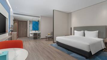 A modern hotel room with a large bed, a desk with a chair, a small table with a plant, and a bathroom visible through an open door.