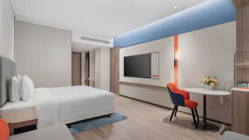 A modern hotel room with a bed, a desk, a chair, a TV, and a small table with a vase of flowers.