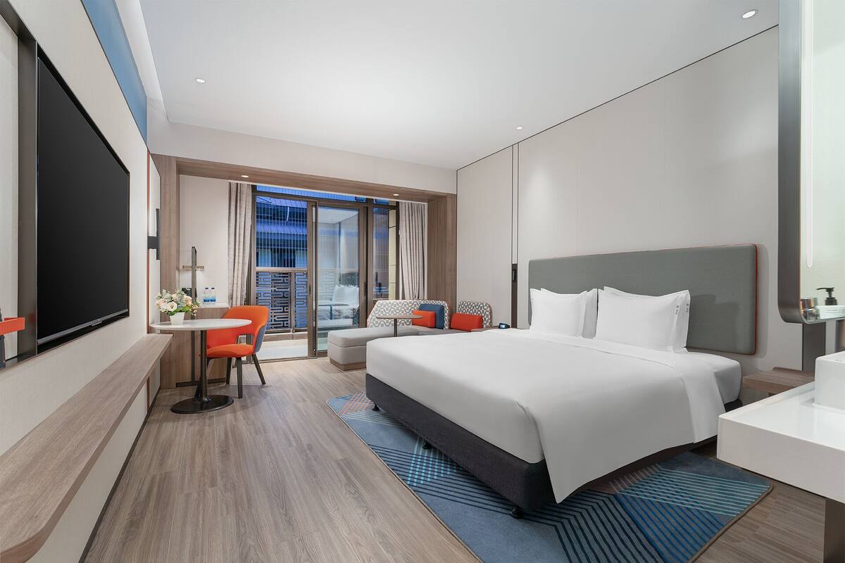 A modern hotel room with a large bed, a desk with a flat-screen TV, a seating area with chairs, and a view of the city through the balcony doors.