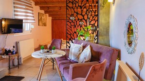 Un salón acogedor con techo de madera, un televisor montado en la pared, un sofá con cojines, una pequeña mesa redonda y un separador decorativo con plantas.