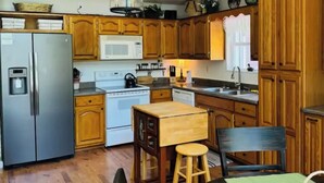 A compact kitchen with wooden cabinets, a stainless steel refrigerator, a microwave, and a sink. There is a wooden table with stools and a hanging light fixture above. - Relax & enjoy the river view, 5 min to Huntsville (Laceys Spring)