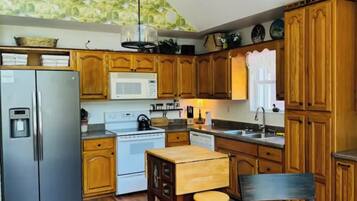 A compact kitchen with wooden cabinets, a stainless steel refrigerator, a microwave, and a sink. There is a wooden table with stools and a hanging light fixture above.
