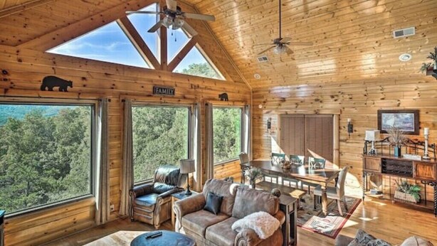 Un'ampia baita di legno con zona pranzo, soggiorno dotato di divano e poltrone, e ampie finestre che si affacciano su una foresta.