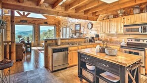 Interno rustico di una baita di legno con cucina, zona pranzo e un'ampia finestra vista su un paesaggio montuoso.