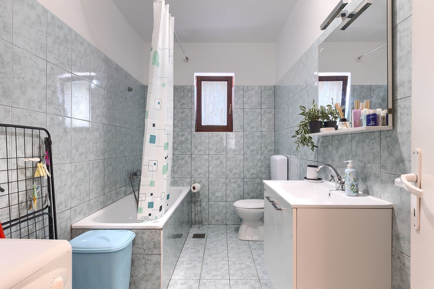A bathroom with a bathtub, toilet, and sink. There is a window above the toilet and a shelf with toiletries above the sink.