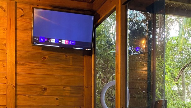 Una cabaña de madera con un jacuzzi, un televisor en la pared y un ventanal de vidrio que da a una terraza con follaje.