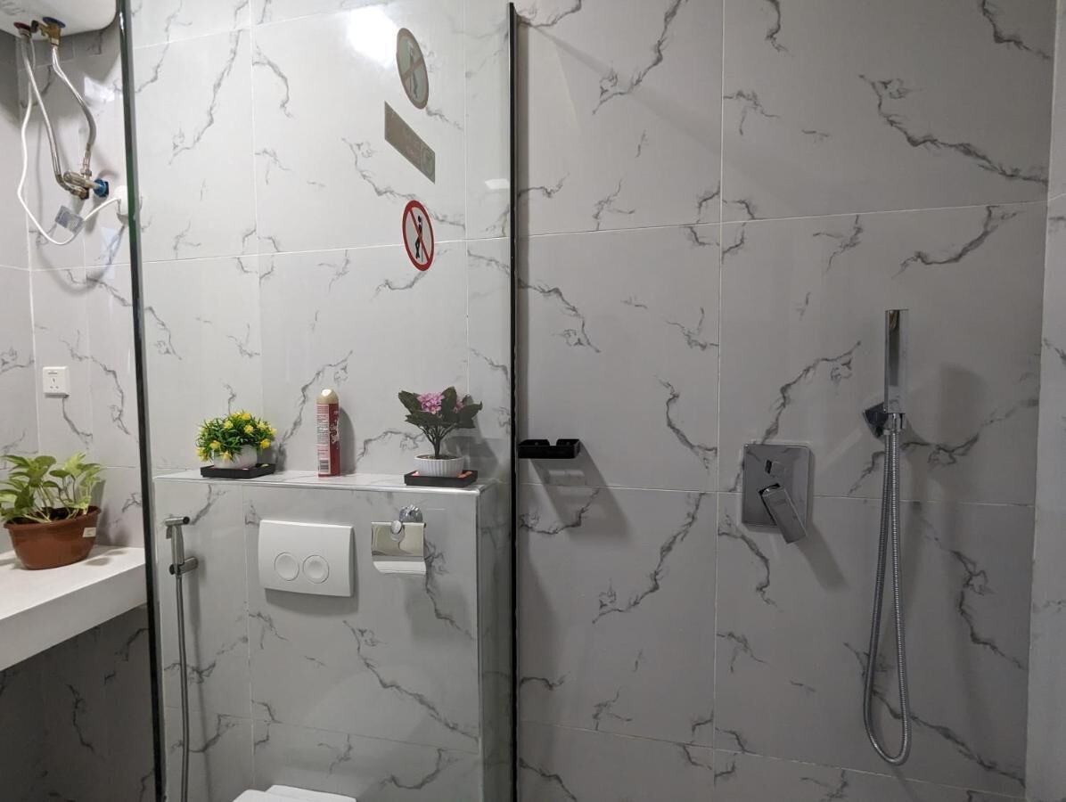 A bathroom with a glass shower enclosure, marble tiles, a toilet, a sink with a soap dispenser, and a potted plant.
