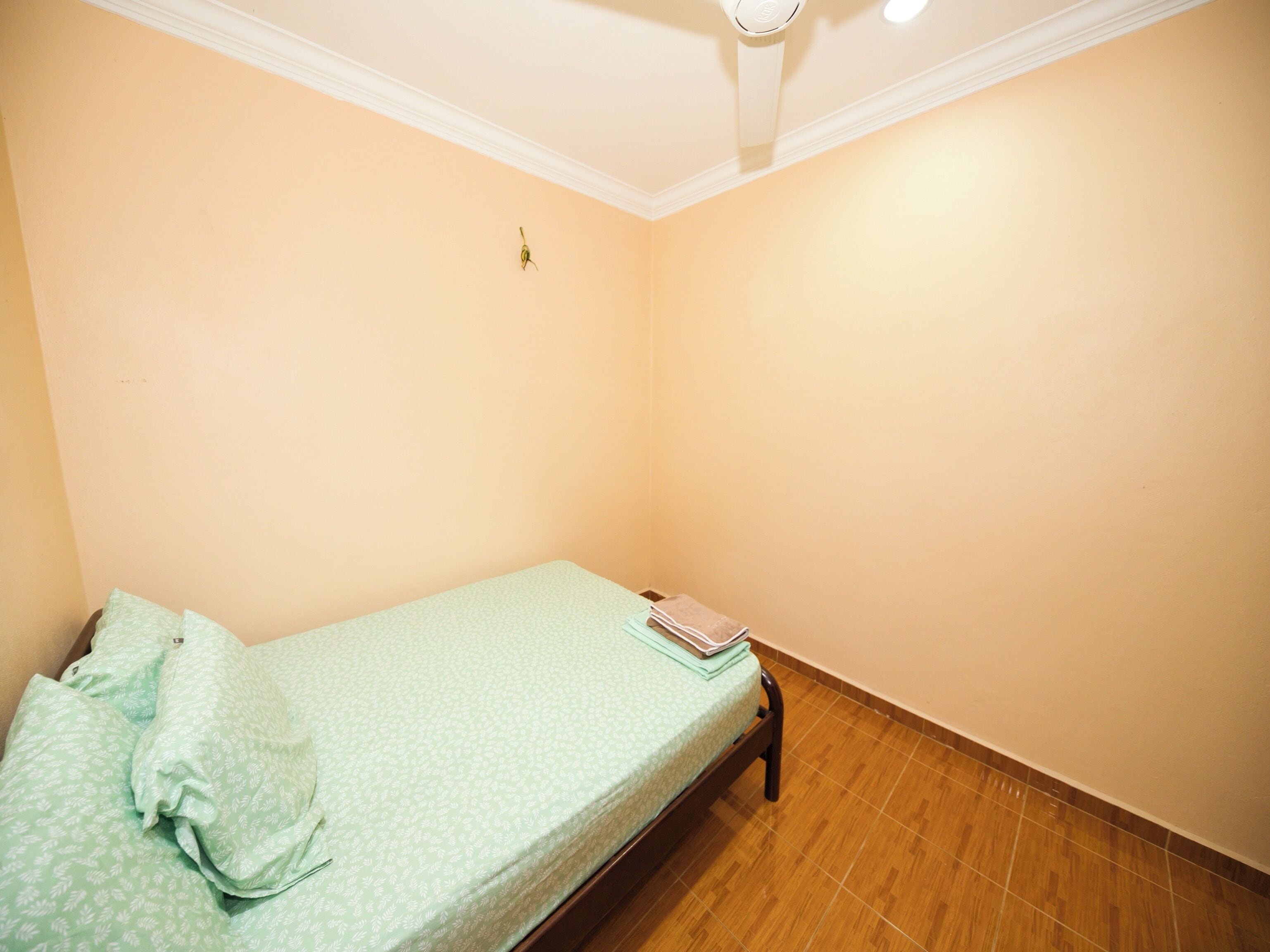 A single bed with a green patterned bedspread, a folded towel, and a book on the bed. The room has peach-colored walls and a wooden floor.