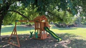 A wooden play structure with a slide and swings, surrounded by trees.