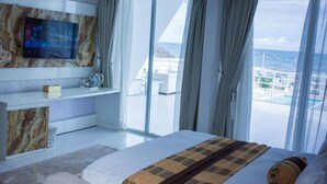 A modern hotel room with a large bed, a flat-screen TV mounted on a marble wall, and a balcony with a view of the ocean. - WHITE DECK BEACH RESORT (Lekki)