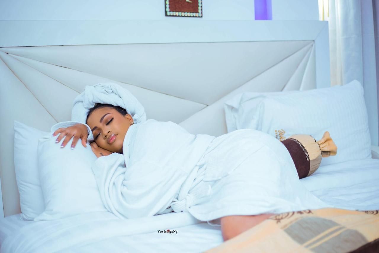 A person lying in bed with white bedding and a headscarf, in a room with a white headboard and a framed picture on the wall.